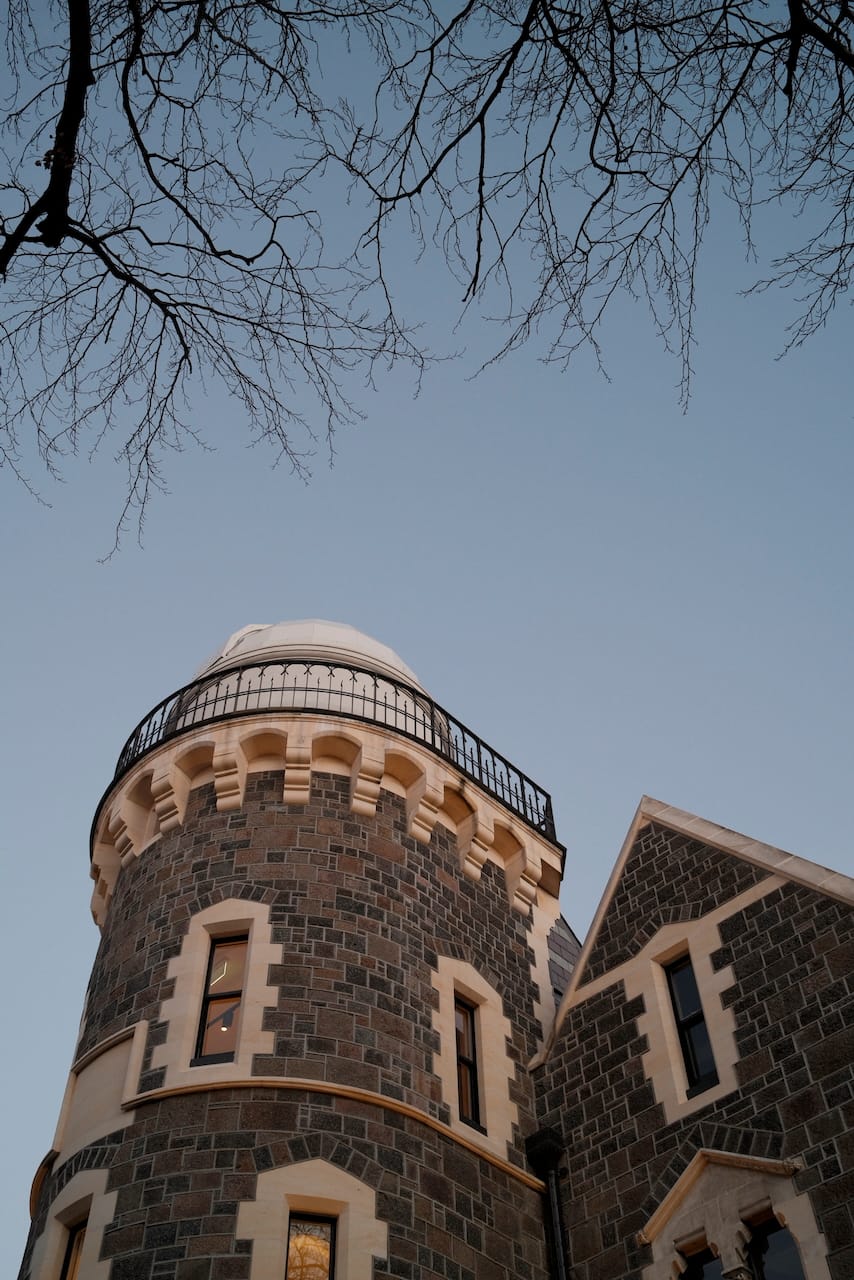 A large stone masonry building in the foreground, being looked up at, toned warm and orange by the last of the sunlight. The tower has a dome at the top for a telescope. Above it in the frame, a dusky sky and leafless branches.