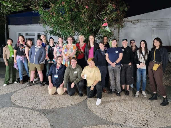 A group of people posing for a photo. They're standing outdoors at night, on a tile floor. There are about 20 of them.