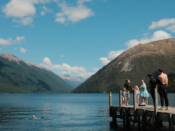 An alpine lake in summer, dramatic mountains in the distance descending into the water. A child is floating on their back in the water as their family look on from a small wharf.